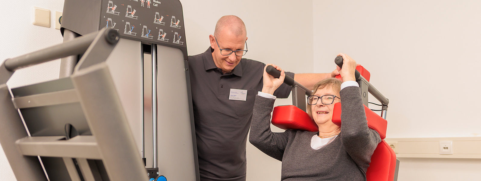 Ältere Frau sitzt an einem Krafttrainingsgerät und drückt die Griffe nach oben, ein Mann steht daneben und beobachtet sie