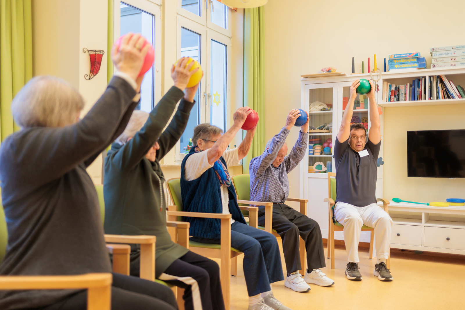 Fünf ältere Menschen sitzen in einem Wohnzimmer auf Stühlen und heben bunte Bälle über den Kopf
